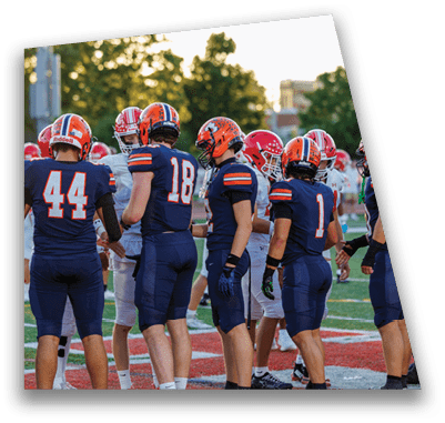 A person in football gear and helmet is crouched on a field with other players lined up in the background during a game. Description generated by AI