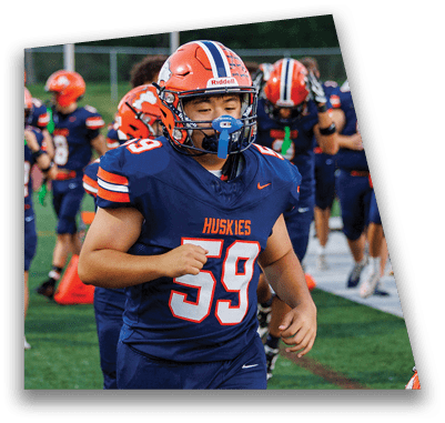 A person in football gear and helmet is crouched on a field with other players lined up in the background during a game. Description generated by AI