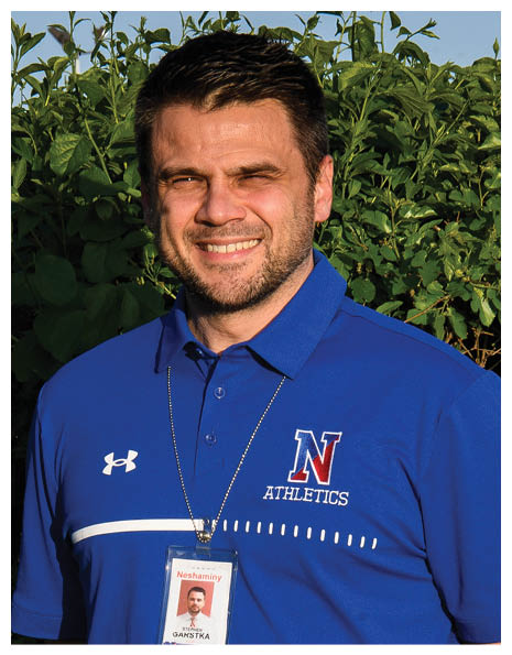 A person wearing a blue polo shirt with an athletics logo and a lanyard with an ID badge is standing outdoors in front of green foliage, smiling. Description generated by AI