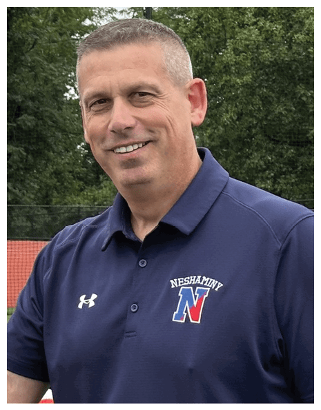 A person wearing a navy blue polo shirt with the word “Neshaminy" and a large "N" logo stands outdoors in front of trees and a fence. Description generated by AI