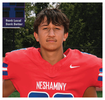 A person with short wavy hair wearing a red Neshaminy football jersey holding a red and white helmet stands on a football field with a scoreboard in the background. Description generated by AI