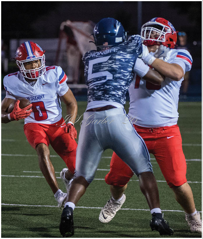 Three people in football uniforms are engaged in a play on a field, with one person holding the ball and another blocking an opponent. Description generated by AI