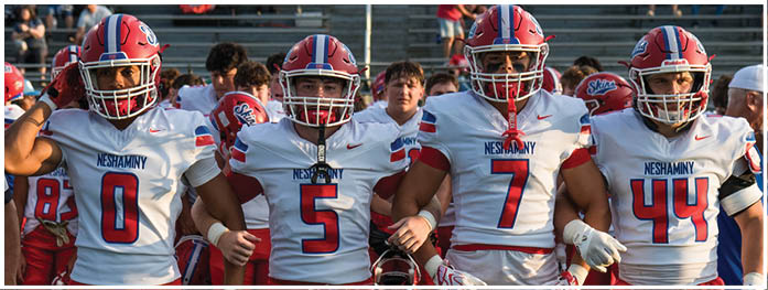 Four people in football uniforms with helmets and gloves stand side by side on a field with stadium seating in the background. Description generated by AI