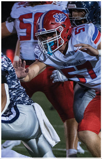People wearing football uniforms are actively engaged in a play on a field, with one person in a dark blue and white uniform running while others in red and white uniforms attempt to catch them. Description generated by AI