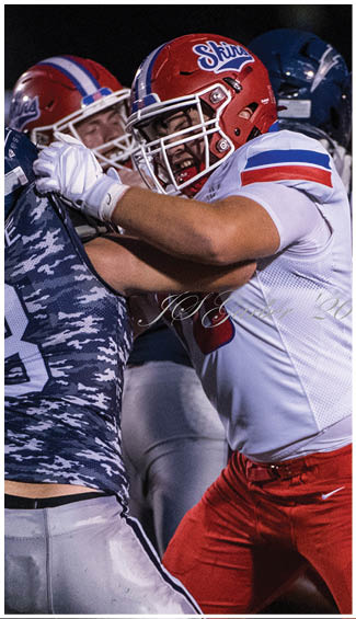 Two people in football uniforms are engaged in a physical play during a game, with one wearing a camouflage-patterned jersey and the other in a white and red uniform. Description generated by AI