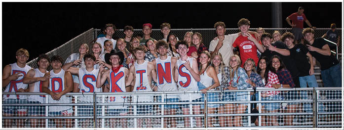 A group of people at a nighttime event stand behind a metal fence, with several wearing white shirts painted with red letters spelling “REDSKINS" and others dressed in casual clothing. Description generated by AI