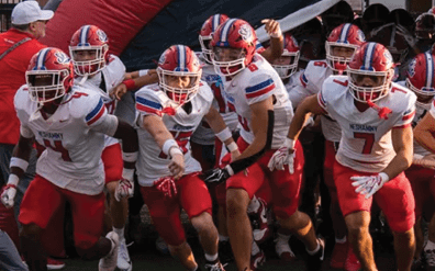 A group of football players in red and white uniforms with helmets are running out onto a field from a tunnel, accompanied by a person in a red vest and cap. Description generated by AI