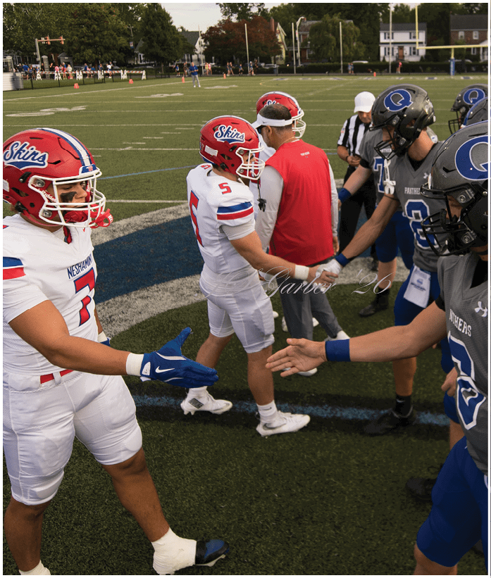 Three people in football uniforms are engaged in a play on a field, with one person holding the ball and another blocking an opponent. Description generated by AI