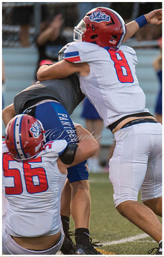 People wearing football uniforms are actively engaged in a play on a field, with one person in a dark blue and white uniform running while others in red and white uniforms attempt to catch them. Description generated by AI
