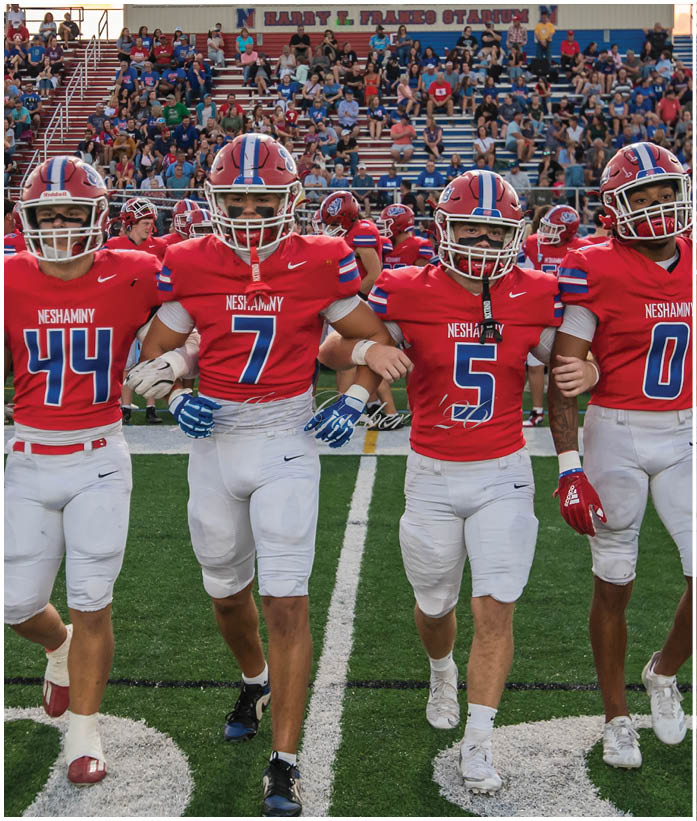 Three people in football uniforms are engaged in a play on a field, with one person holding the ball and another blocking an opponent. Description generated by AI