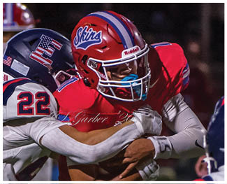 People wearing football uniforms are engaged in a physical play on a field during a game at night. Description generated by AI