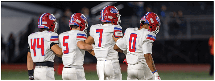 Four people in football uniforms with helmets and gloves stand side by side on a field with stadium seating in the background. Description generated by AI