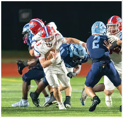 Several people wearing football helmets and uniforms are actively engaged in a play on a football field at night. Description generated by AI