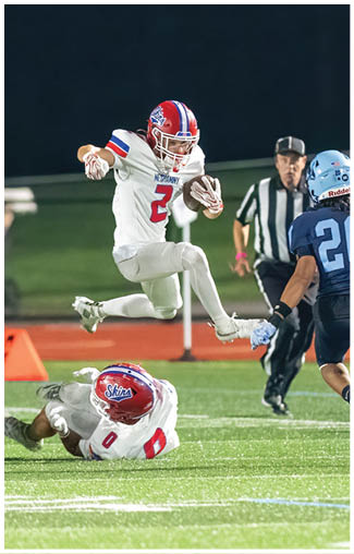 People wearing football uniforms are actively engaged in a play on a field, with one person in a dark blue and white uniform running while others in red and white uniforms attempt to catch them. Description generated by AI