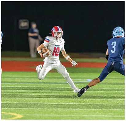 Several people wearing football helmets and uniforms are actively engaged in a play on a football field at night. Description generated by AI