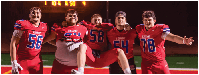 Four people in football uniforms with helmets and gloves stand side by side on a field with stadium seating in the background. Description generated by AI