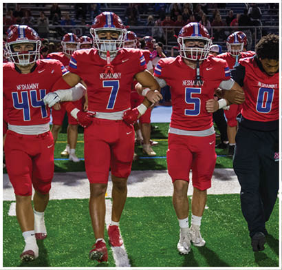Several people wearing football helmets and uniforms are actively engaged in a play on a football field at night. Description generated by AI