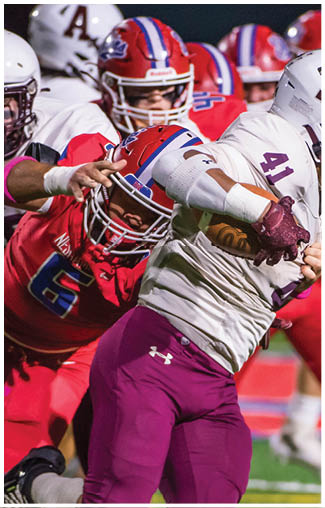 People wearing football uniforms are actively engaged in a play on a field, with one person in a dark blue and white uniform running while others in red and white uniforms attempt to catch them. Description generated by AI