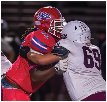 Several people wearing football helmets and uniforms are actively engaged in a play on a football field at night. Description generated by AI