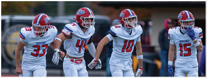 Four people in football uniforms with helmets and gloves stand side by side on a field with stadium seating in the background. Description generated by AI