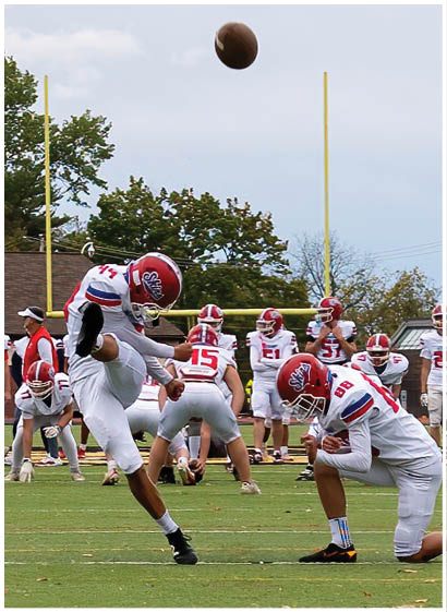 A group of people wearing red and white football uniforms are actively playing on a field, with one person holding the ball and running while others surround them. Description generated by AI