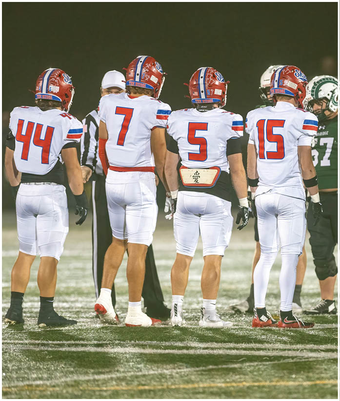 Three people in football uniforms are engaged in a play on a field, with one person holding the ball and another blocking an opponent. Description generated by AI