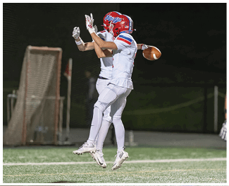 People wearing football uniforms are engaged in a physical play on a field during a game at night. Description generated by AI