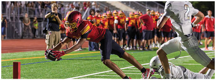 A football player in a red and yellow jersey is diving for a ball. Description generated by AI