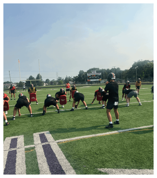 A football game is in progress with players on the field. One player is throwing a football while another player is trying to catch it. The players are wearing uniforms and are actively engaged in the game. AI generated content