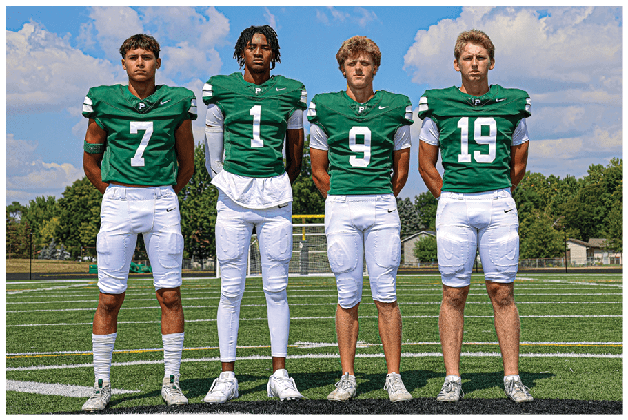 Four adults wearing green football jerseys and white pants stand side by side on a football field under a partly cloudy sky. Description generated by AI