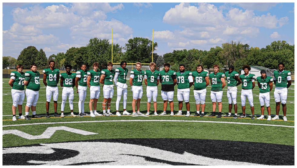 A group of people wearing green football jerseys and white pants stand in a line on a football field under a partly cloudy sky. Description generated by AI