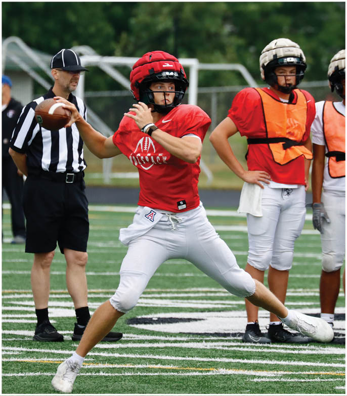 An adult in a red football uniform is preparing to throw a football while three other adults in helmets and orange practice vests stand on a field with a referee nearby. Description generated by AI