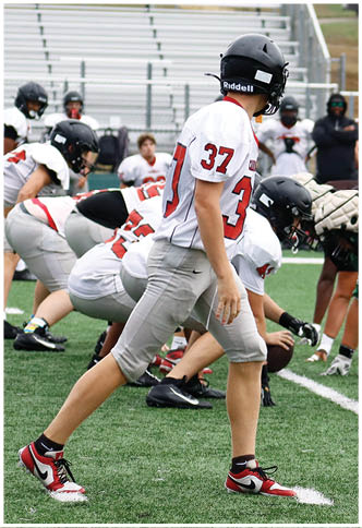 Players in football uniforms are lined up on a field preparing for a play with one player standing and others crouched at the line of scrimmage. Description generated by AI