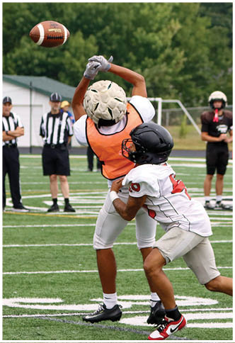 Two people in football gear are on a field with one person in an orange vest reaching up to catch a ball while the other person in a white jersey and black helmet attempts to block or tackle. Description generated by AI