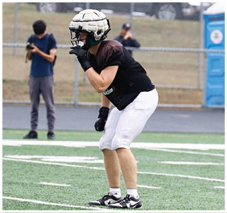 A person wearing a padded helmet, black sports jersey, white pants, and gloves is crouched in an athletic stance on a field with a blurred background of two people and a fence. Description generated by AI