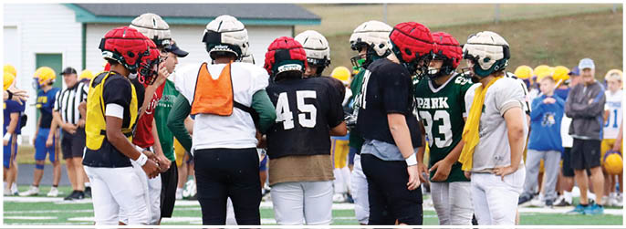 A group of people wearing football helmets and jerseys are standing together on a field while others watch in the background. Description generated by AI