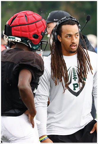 A person with long dreadlocks wearing a white long-sleeve shirt with a football logo is standing and talking to another person in football gear with a red helmet. Description generated by AI