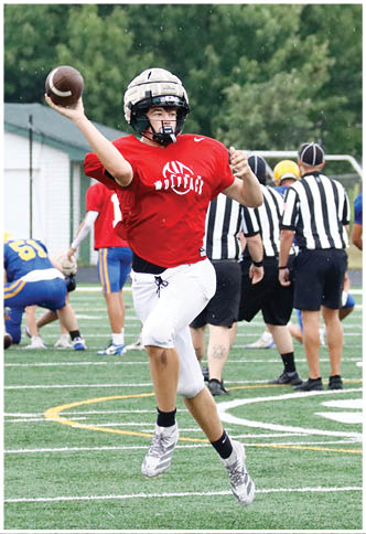 A person wearing a red sports jersey and white pants is jumping and throwing a football on a green field with other players and referees in the background. Description generated by AI
