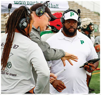Two adults are engaged in a conversation, one wearing a white polo shirt with a green “P" and a headset, while the other gestures with their hand. Description generated by AI