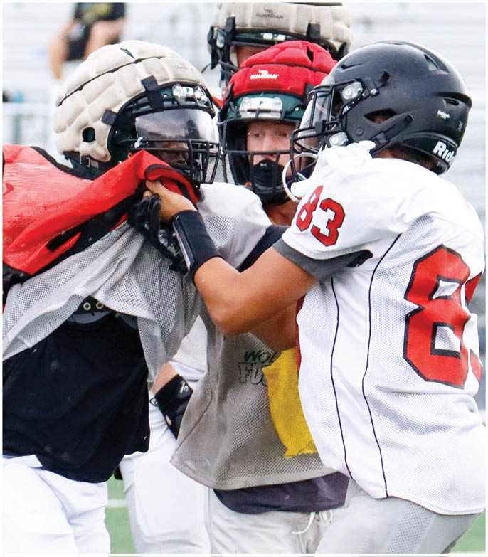 Two people wearing football helmets and jerseys are engaged in a physical drill on a field while a third person watches closely. Description generated by AI