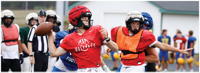 A person wearing a red helmet and red shirt is throwing a football while being pursued by others in protective gear on a sports field. Description generated by AI
