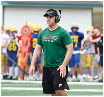 A person wearing a green Park P Football shirt, a black visor, and a headset stands on a sports field with blurred players and equipment in the background. Description generated by AI