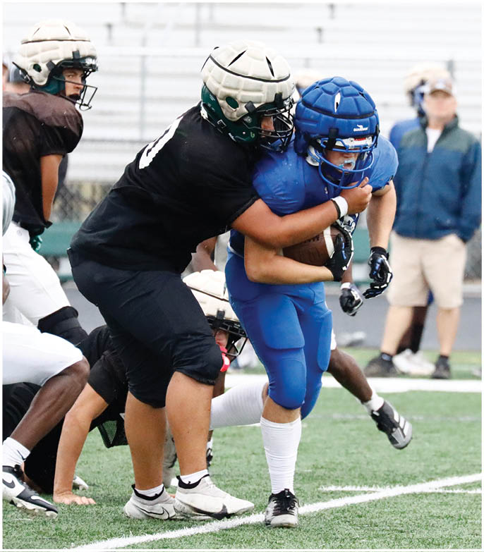 Two people in football gear are engaged in a tackle on a field with others and a coach in the background. Description generated by AI