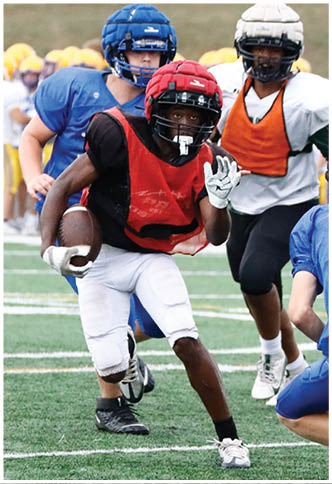 A person wearing a blue football jersey with the number 36 and a black helmet is crouched on a field while other people in sports attire stand nearby. Description generated by AI