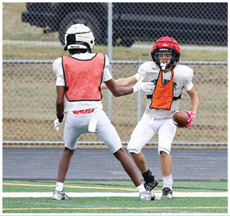 Two people wearing football helmets and orange practice vests are engaged in a play on a grassy field with a chain-link fence in the background. Description generated by AI