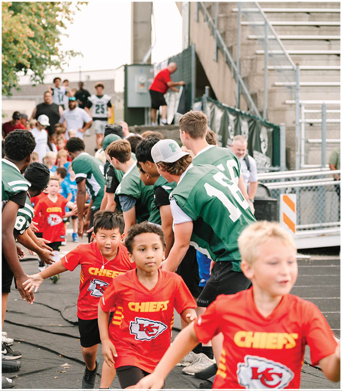 Children wearing red Kansas City Chiefs shirts run through a line of people in green sports jerseys giving high-fives on an outdoor track with a tree and spectators in the background. Description generated by AI