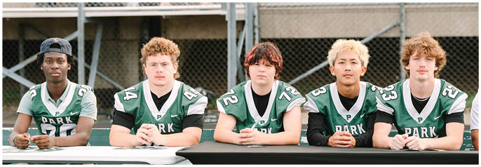 Five people wearing green and white football jerseys are sitting side by side at a table in front of a chain-link fence. Description generated by AI