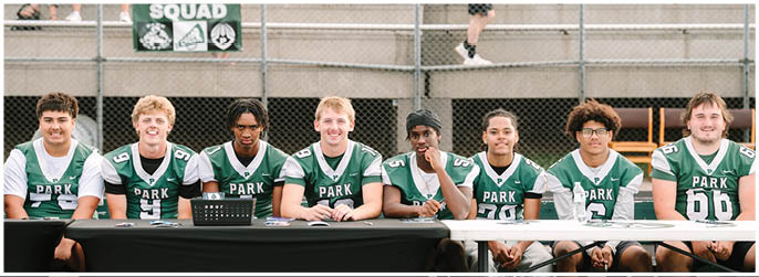 A group of people wearing green and white sports jerseys are sitting in front of bleachers with a “Cheer Squad" banner hanging behind them. Description generated by AI
