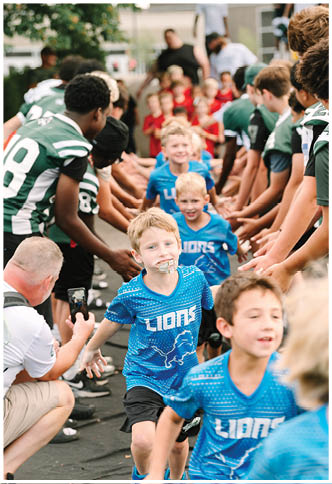 Children in blue shirts are smiling and walking through a tunnel formed by adults and other children giving high fives outdoors. Description generated by AI