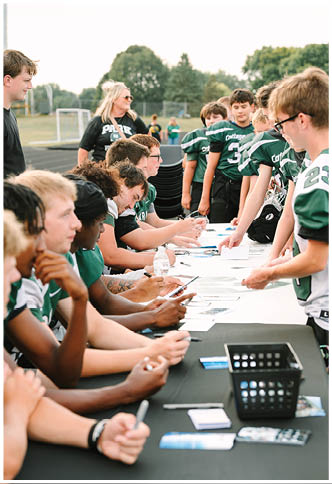 Children in green football uniforms are gathered outdoors with adults and trees in the background. Description generated by AI